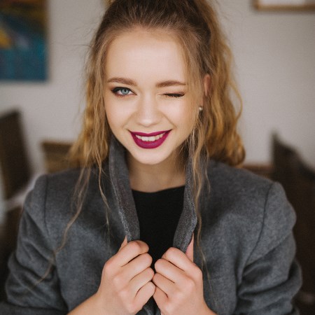 Blondynka z pofalowanymi włosami, ustami pomalowanymi na bordowo i w szarej marynarce