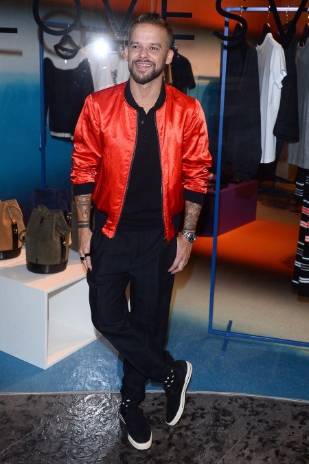 Michał Piróg w pomarańczowej kurtce bomber, czarnej koszuli i eleganckich spodniach w kant oraz trampkach.