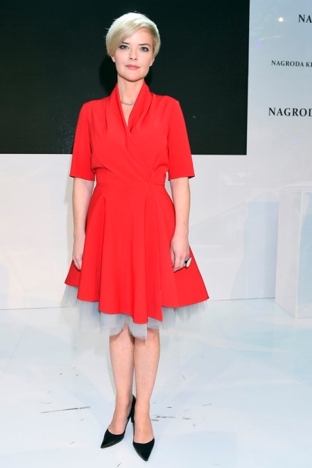 Monika Zamachowska ubrana w czerwoną sukienkę rozkloszowaną, delikatny, srebrny naszyjnik i czarne szpilki.